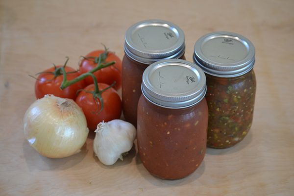 Jars of Millcreek Miracles homemade salsa beside fresh tomatoes, an onion and garlic
